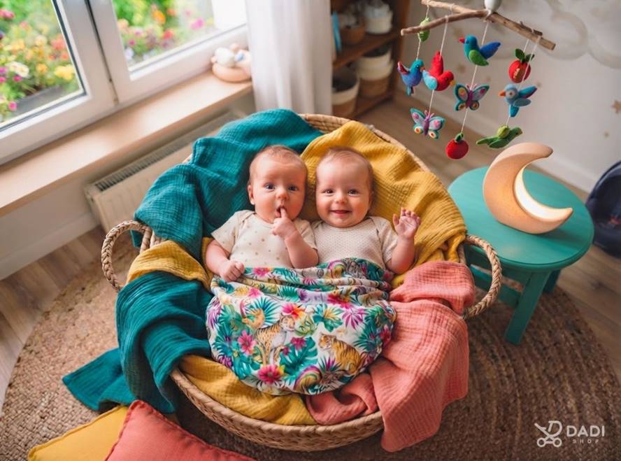 Dwoje niemowląt leży obok siebie w wiklinowym koszu w przytulnym pokoju dziecięcym. 